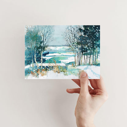 Hand holding a greeting card featuring a watercolor winter landscape of snowy fields, bare trees, and evergreens in shades of teal, blue, and white, with splattered paint details suggesting falling snow.