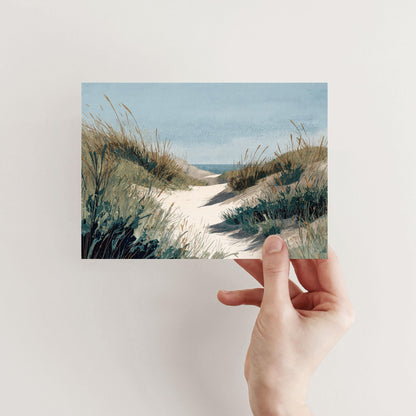 Hand holding a greeting card featuring a watercolor illustration of a sandy path through grassy dunes leading toward the ocean under a clear blue sky.