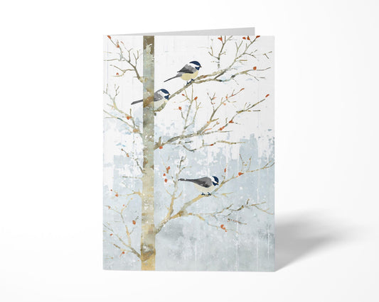 Folded greeting card featuring a watercolor-style illustration of three chickadees perched on bare winter branches. Soft gray-blue snowy background with scattered orange berries creates a calm, wintry feel. Card stands upright on a white surface with a faint shadow.