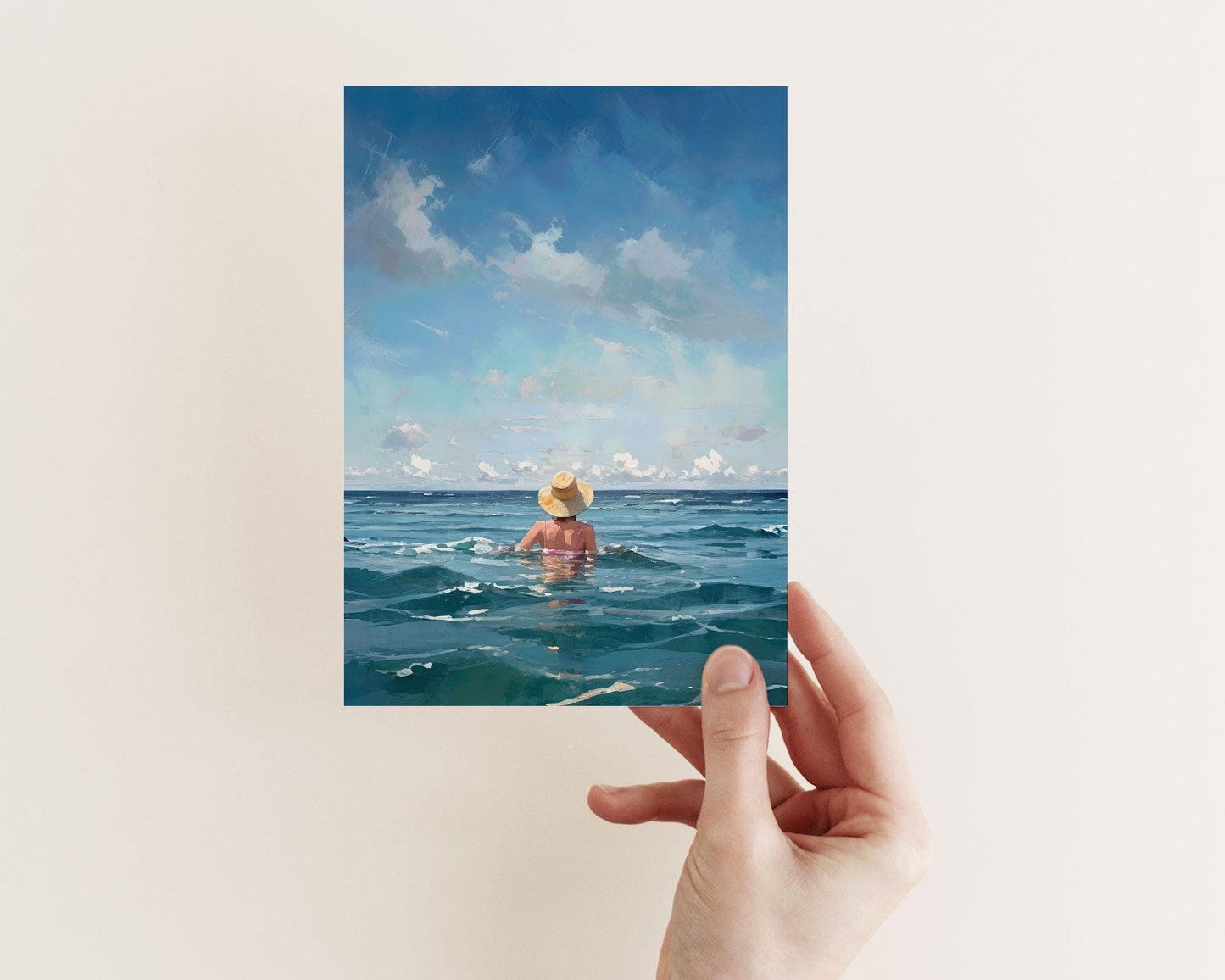 Hand holding a card featuring a painting of a woman in a sunhat facing the ocean, surrounded by rippling teal water and a sky scattered with clouds.