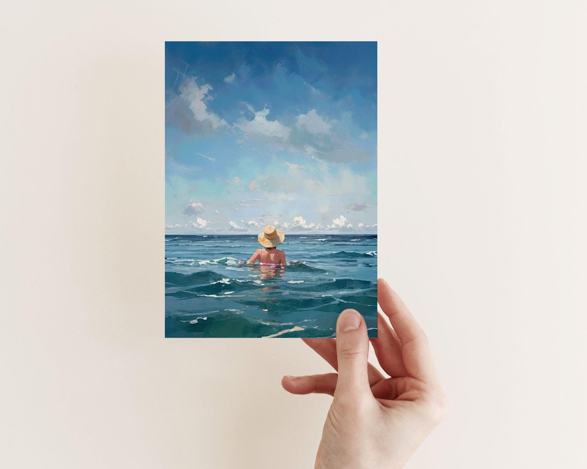 Hand holding a card featuring a painting of a woman in a sunhat facing the ocean, surrounded by rippling teal water and a sky scattered with clouds.