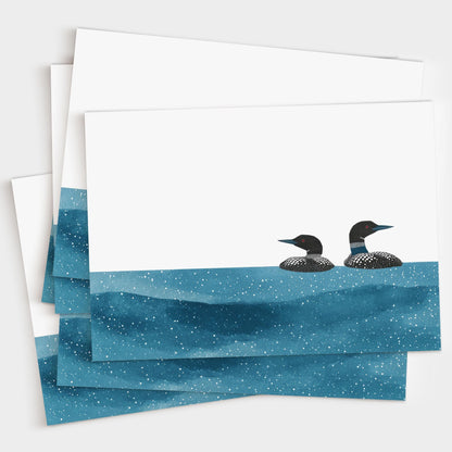 Set of minimalist greeting cards featuring two loons floating on deep blue, star-speckled water against a crisp white background, fanned out to display multiple copies.