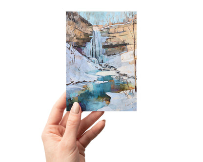 Hand holding a greeting card featuring a watercolor-style painting of a winter waterfall scene. The artwork shows a cascade flowing over rocky cliffs into a turquoise pool surrounded by snow and ice, with bare trees silhouetted against a pale blue sky.