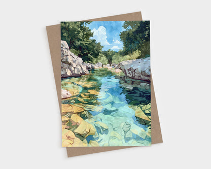 Greeting card featuring a watercolor-style painting of a crystal-clear river with golden rocks beneath the surface, bordered by white stone banks and lush green trees under a bright blue sky with clouds. Card is paired with a kraft envelope.