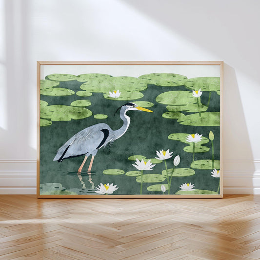 Framed art print of a blue heron wading through a pond filled with green lily pads and white lotus flowers, illustrated in a clean, modern style with textured watercolor effects. The print leans against a white wall on a light wood herringbone floor, with natural light casting soft shadows.