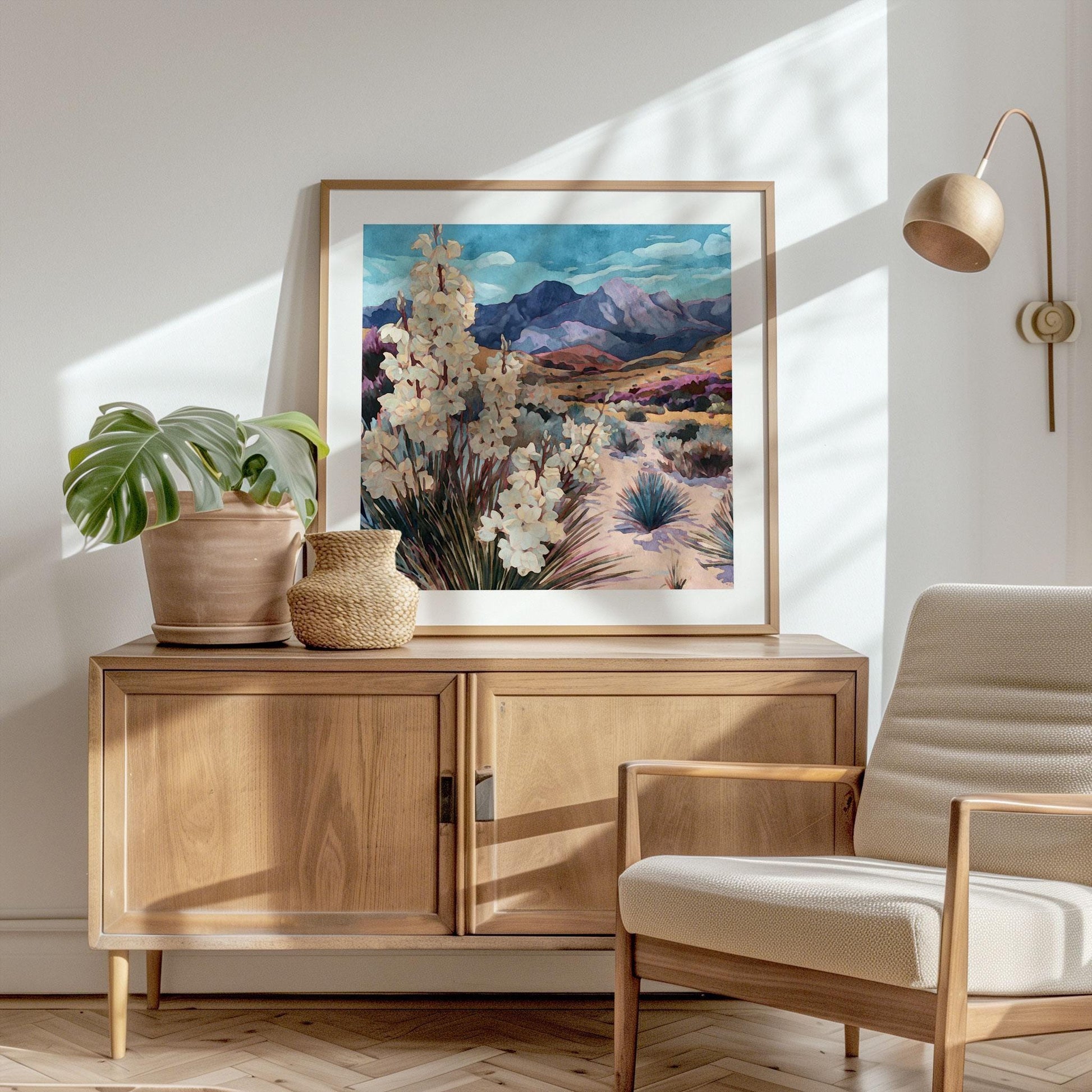 Framed desert landscape painting featuring blooming yucca plants, sandy hills, and purple-hued mountains under a bright blue sky, displayed on a wooden cabinet beside potted plants in a sunlit modern living room.