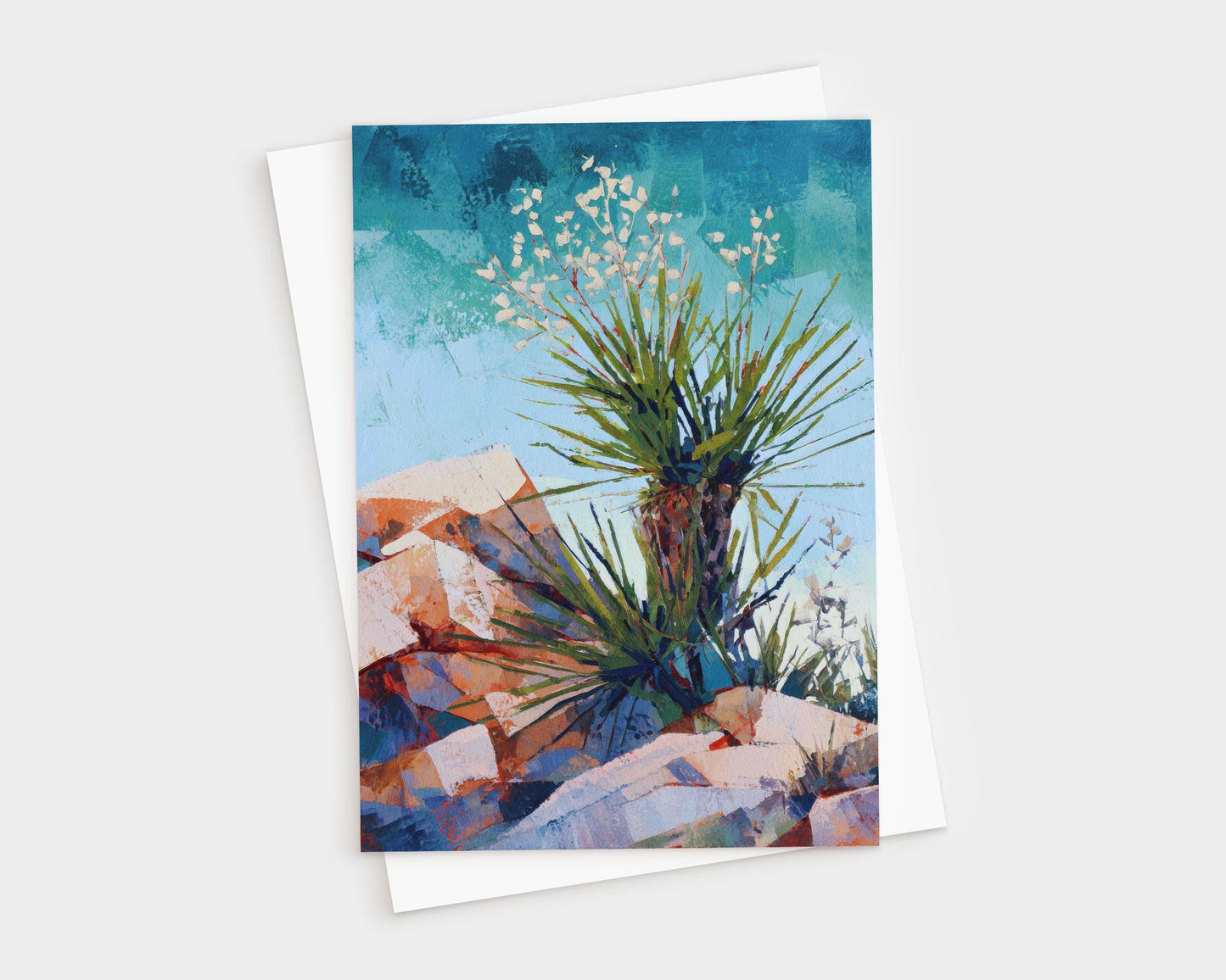 Greeting card featuring a colorful desert landscape painting of a yucca plant with spiky green leaves and small white blossoms, set against a vivid blue sky and warm-toned rocky terrain. Card is shown with a white envelope.