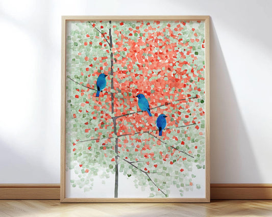 Framed art print of three bright bluebirds perched on thin branches of a maple tree with green and orange-red leaves, painted in a soft, mosaic-like watercolor style. The artwork leans against a white wall on a light wood floor, surrounded by natural sunlight.
