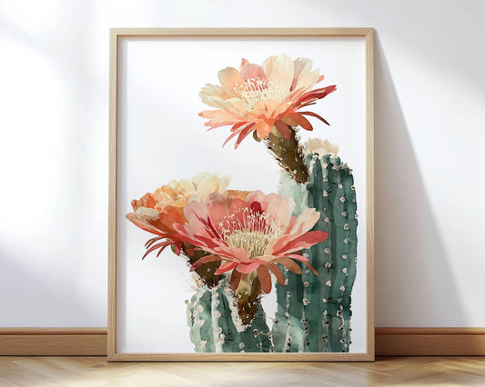 Watercolor art print of a cactus with three large blooming flowers in warm shades of peach, coral, and orange, set against a clean white background and framed in a light wood frame.