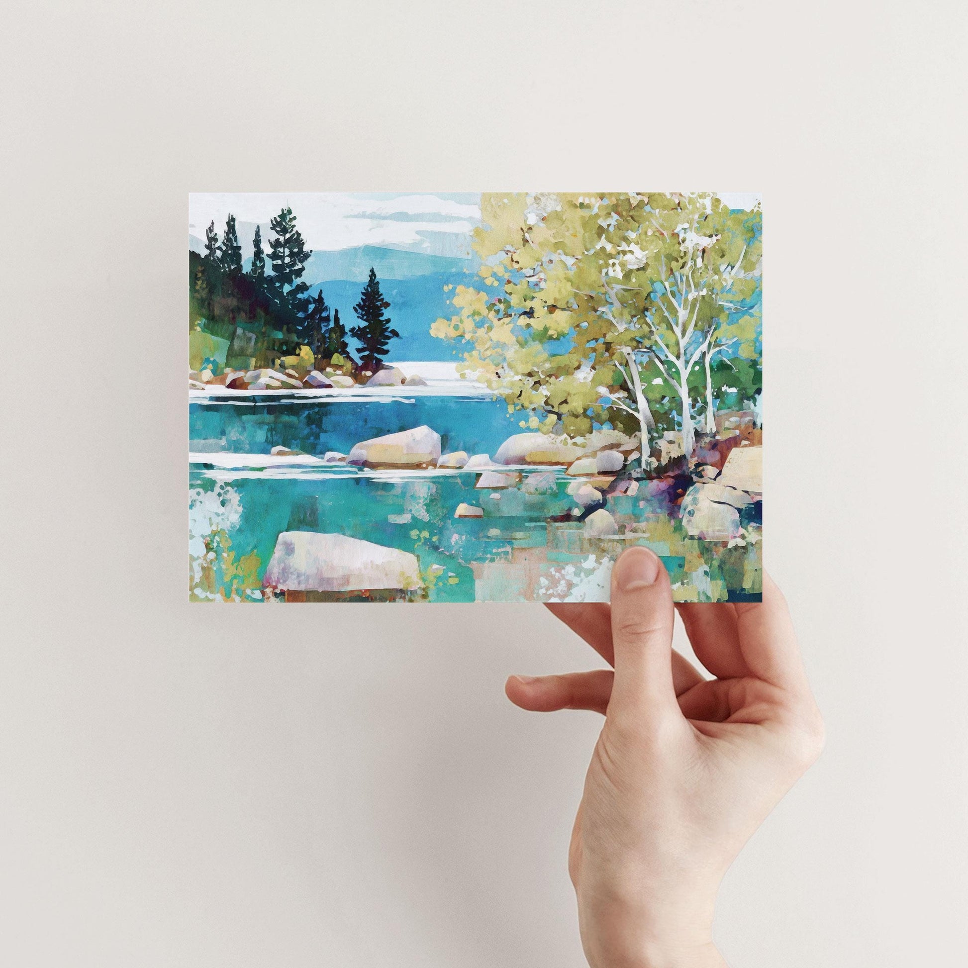 Hand holding a greeting card featuring a colorful impressionist landscape of a lake with turquoise water, smooth rocks, and shoreline trees. Golden-green birches with white trunks frame the right side, while dark evergreen trees and distant blue mountains rise in the background under a soft sky.