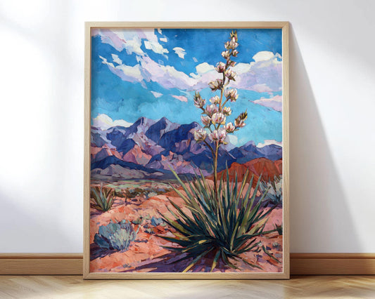 Framed art print of a desert landscape painting featuring a yucca plant with white blooms in the foreground, red sand and sagebrush in the midground, and purple mountain ranges under a vivid blue sky with wispy clouds. The artwork has bold, faceted brushstrokes and a modern impressionist style, displayed in a light wood frame leaning against a white wall on a hardwood floor.