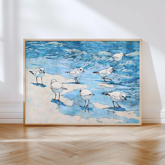 Framed coastal art print depicting a group of small white shorebirds with orange beaks and legs, standing and wading along the shoreline. The scene features soft sandy areas and vibrant blue ocean waves, painted in a fluid, watercolor style with black ink outlines. The artwork rests against a white wall on a light wood herringbone floor, bathed in natural sunlight.
