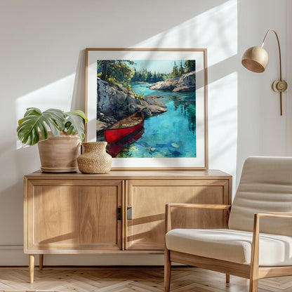Framed art print of a red canoe floating in clear turquoise water, displayed on a wooden sideboard in a bright, modern room. Soft sunlight casts shadows on the wall, highlighting a potted plant, woven vase, and a light wood lounge chair beside the artwork.