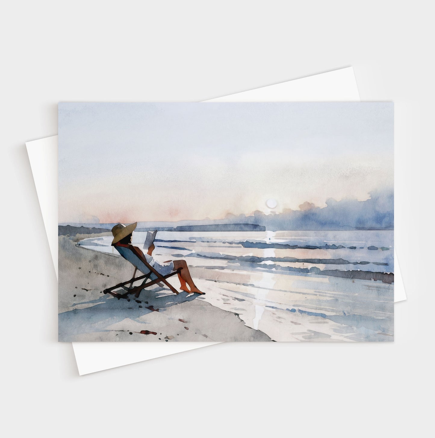 Greeting card with a watercolor-style illustration of a woman lounging in a beach chair reading a book at sunset, with soft waves and pastel sky in the background.
