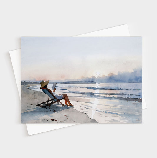 Greeting card with a watercolor-style illustration of a woman lounging in a beach chair reading a book at sunset, with soft waves and pastel sky in the background.