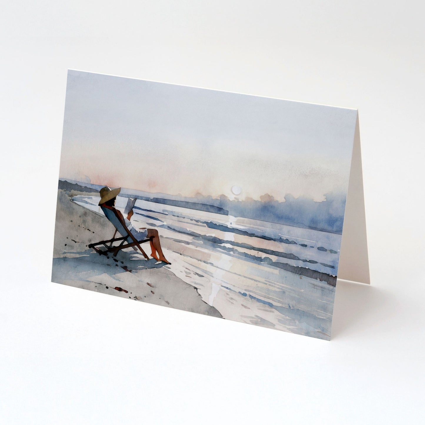 Minimalist greeting card standing open, featuring a watercolor-style illustration of a woman reading in a beach chair at sunset beside calm ocean waves.