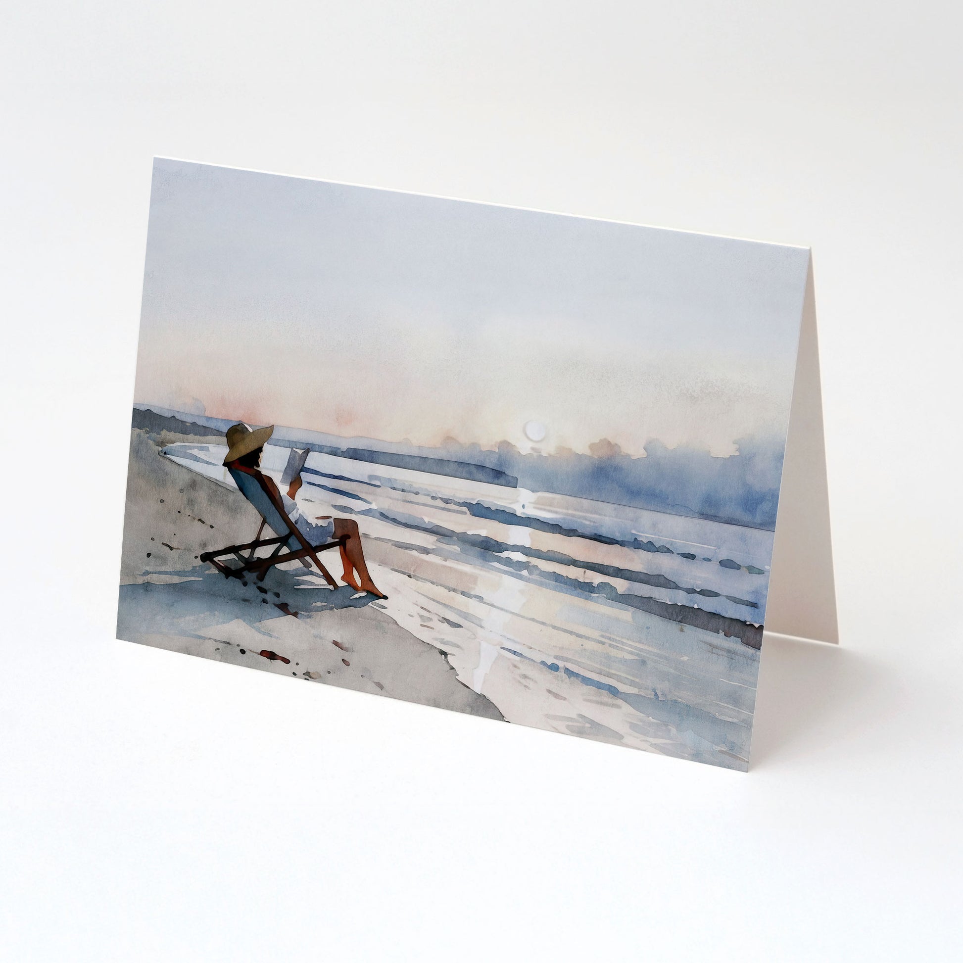 Minimalist greeting card standing open, featuring a watercolor-style illustration of a woman reading in a beach chair at sunset beside calm ocean waves.
