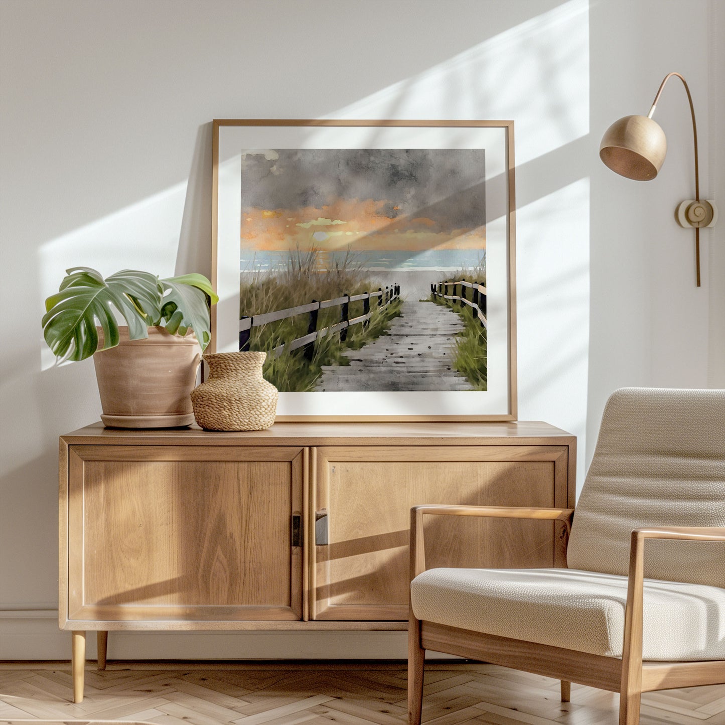 Framed artwork in a natural wood frame, leaning on a wooden console in a bright, sunlit room, depicting a watercolor-style boardwalk leading to the ocean at sunset. The console is styled with a potted plant and a woven basket.