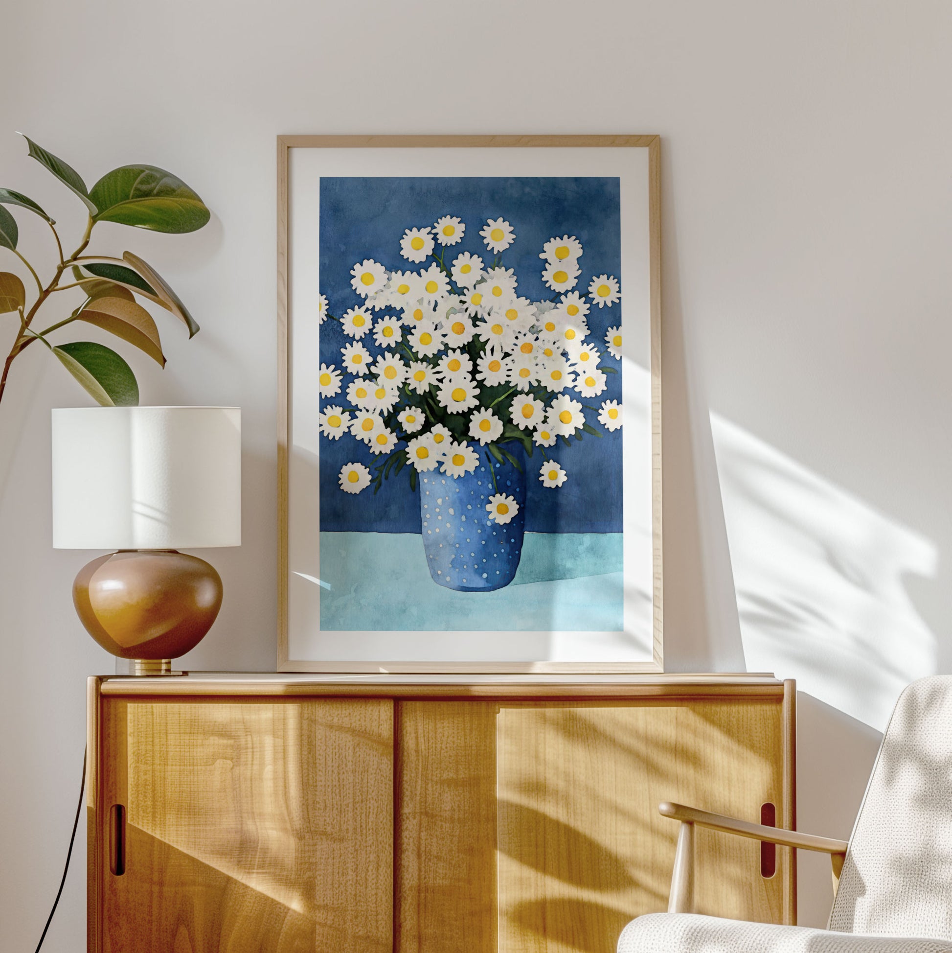 Framed artwork featuring white chamomile flowers with yellow centers in a dotted blue vase leaning on a wooden cabinet, styled with a brown ceramic lamp, a leafy plant, and a beige armchair.