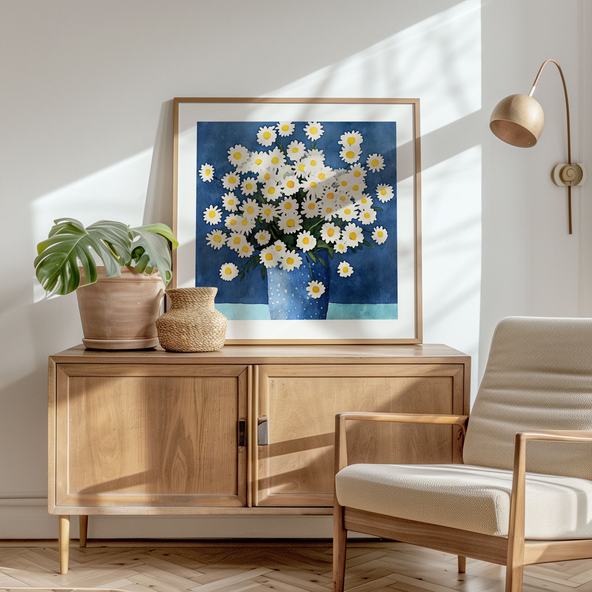 Framed artwork featuring white chamomile flowers with yellow centers in a dotted blue vase leaning on a wooden cabinet, accompanied by a potted monstera plant, a woven basket, and a neutral armchair.