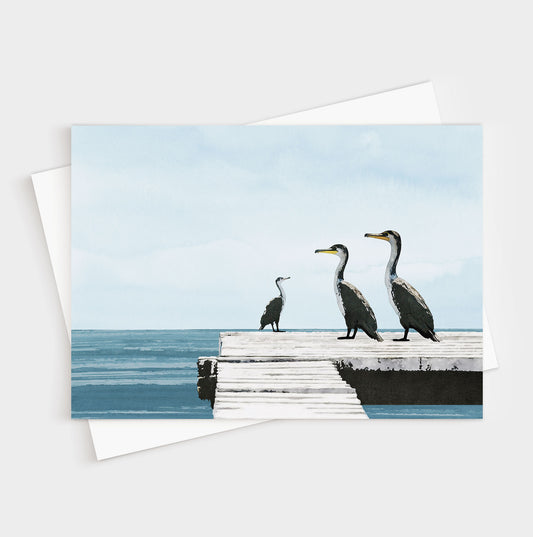 Greeting card with a minimalist illustration of three cormorants standing on a weathered white dock overlooking calm blue ocean water under a pale sky, with a white envelope beneath.