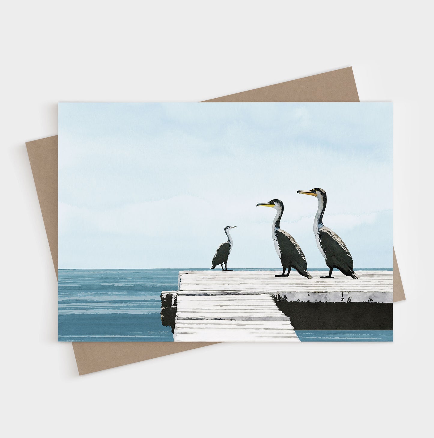 Greeting card with a minimalist illustration of three cormorants standing on a weathered white dock overlooking calm blue ocean water under a pale sky, with a kraft envelope beneath.