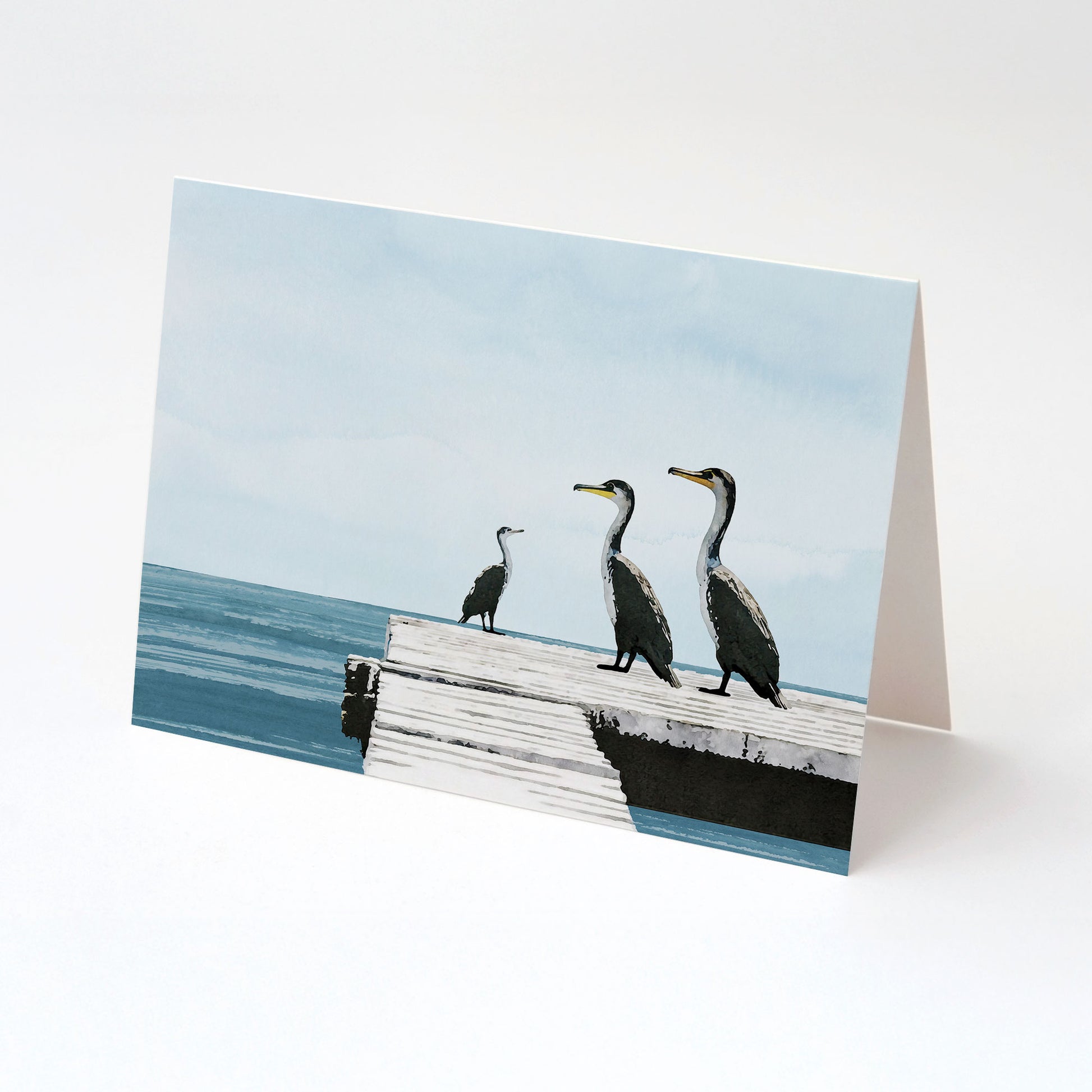 Greeting card standing upright, featuring a minimalist illustration of three cormorants on a white wooden dock by calm blue ocean water, with a soft sky in the background.