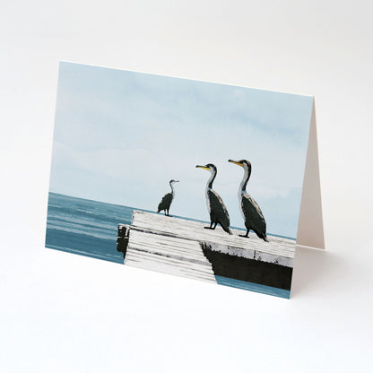 Greeting card standing upright, featuring a minimalist illustration of three cormorants on a white wooden dock by calm blue ocean water, with a soft sky in the background.