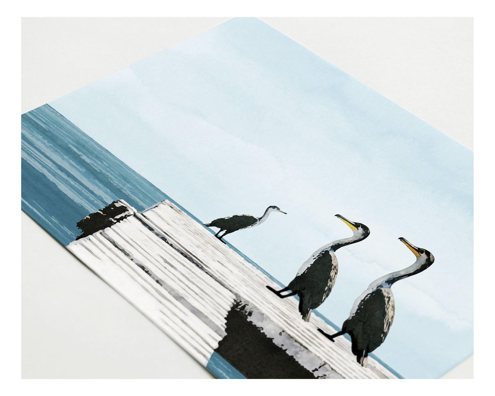 Close-up of a greeting card lying flat, featuring a digital painting of three cormorants standing on a weathered white dock overlooking calm blue ocean water under a pale sky.