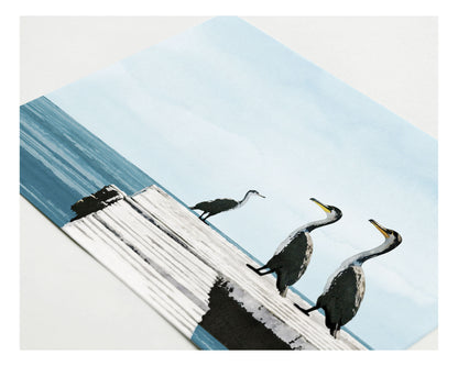 Close-up of a greeting card lying flat, featuring a digital painting of three cormorants standing on a weathered white dock overlooking calm blue ocean water under a pale sky.