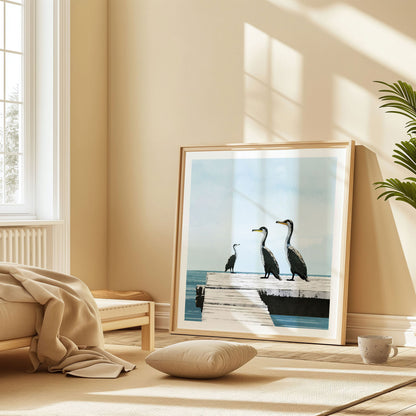 Large framed artwork of three cormorants on a wooden dock rests against a wall in a sunlit room with soft neutral tones, a low bed, cushion, and a potted plant. The ocean scene contrasts with the warm, cozy interior.