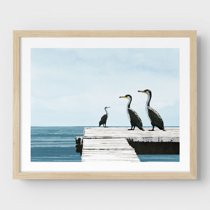Natural wood framed illustration of three cormorants standing on a white wooden dock over a calm blue ocean under a pale sky, with minimalist styling and clean lines against a white background.