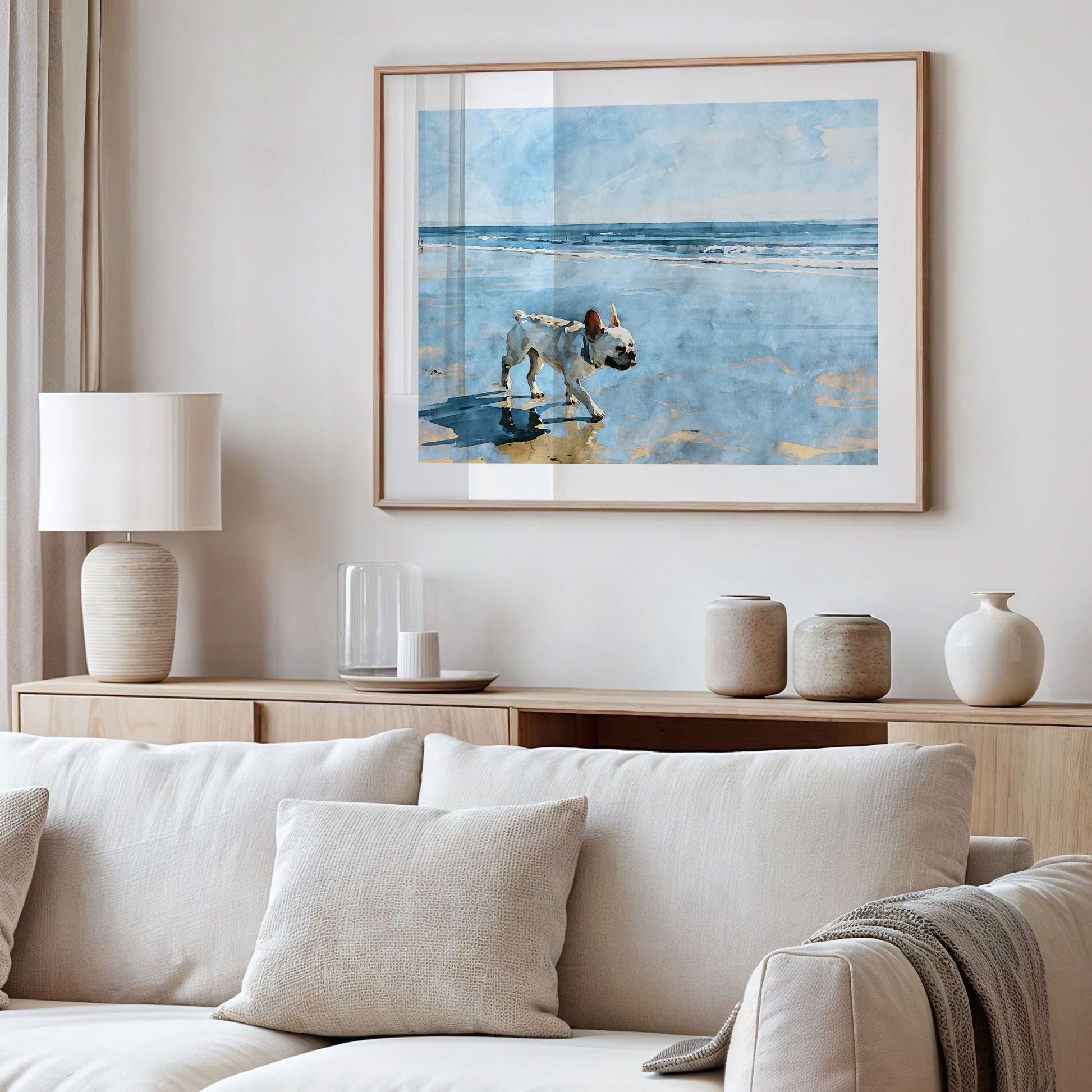Framed cream French Bulldog beach watercolor art displayed in a styled interior with soft blue and sandy neutral tones.