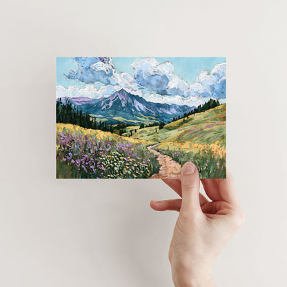 Hand holding a postcard with a mountain landscape and colorful fields.
