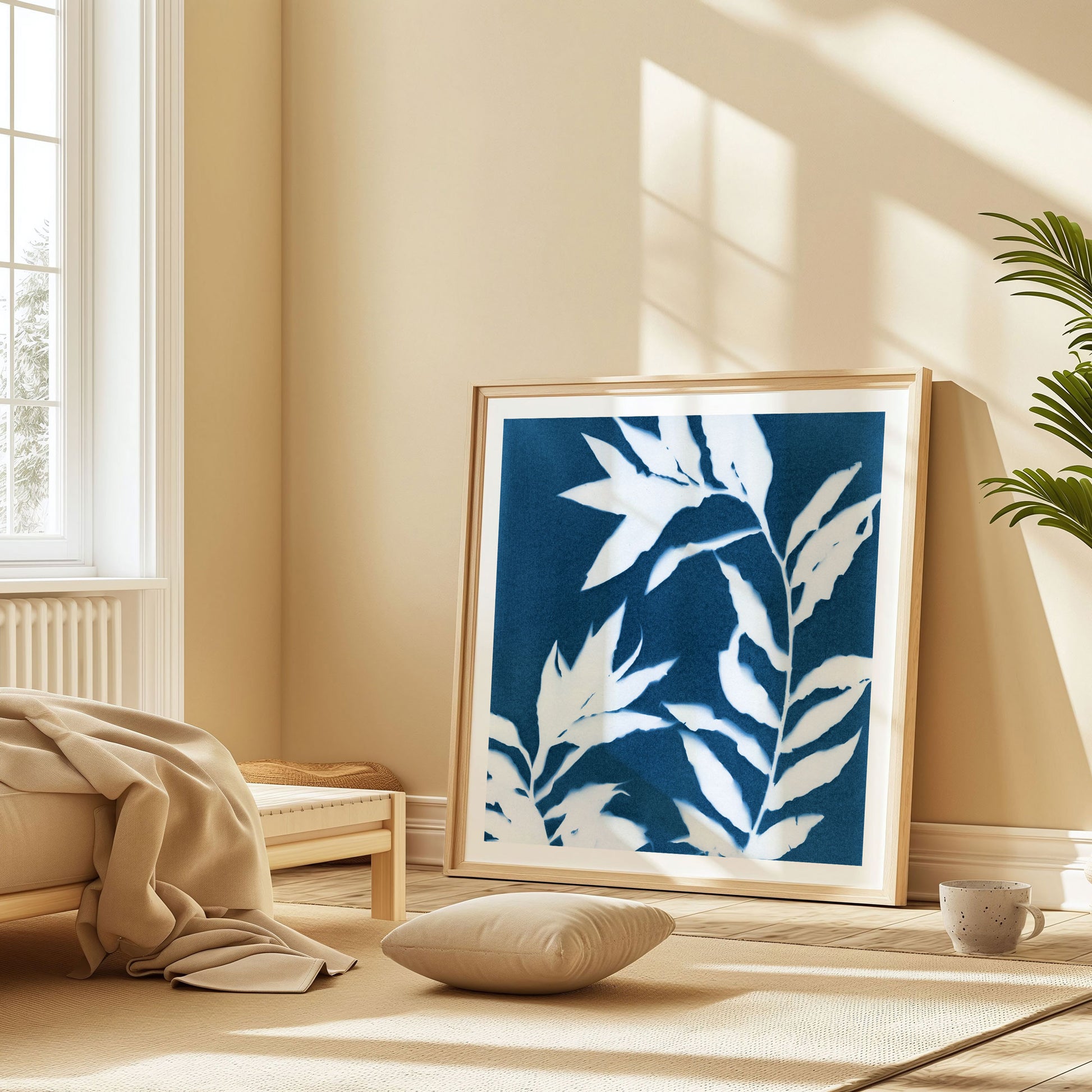 Large framed cyanotype print featuring curved branches with pointed leaves in white against a deep blue background, leaning against a beige wall in a sunlit, minimalist bedroom.