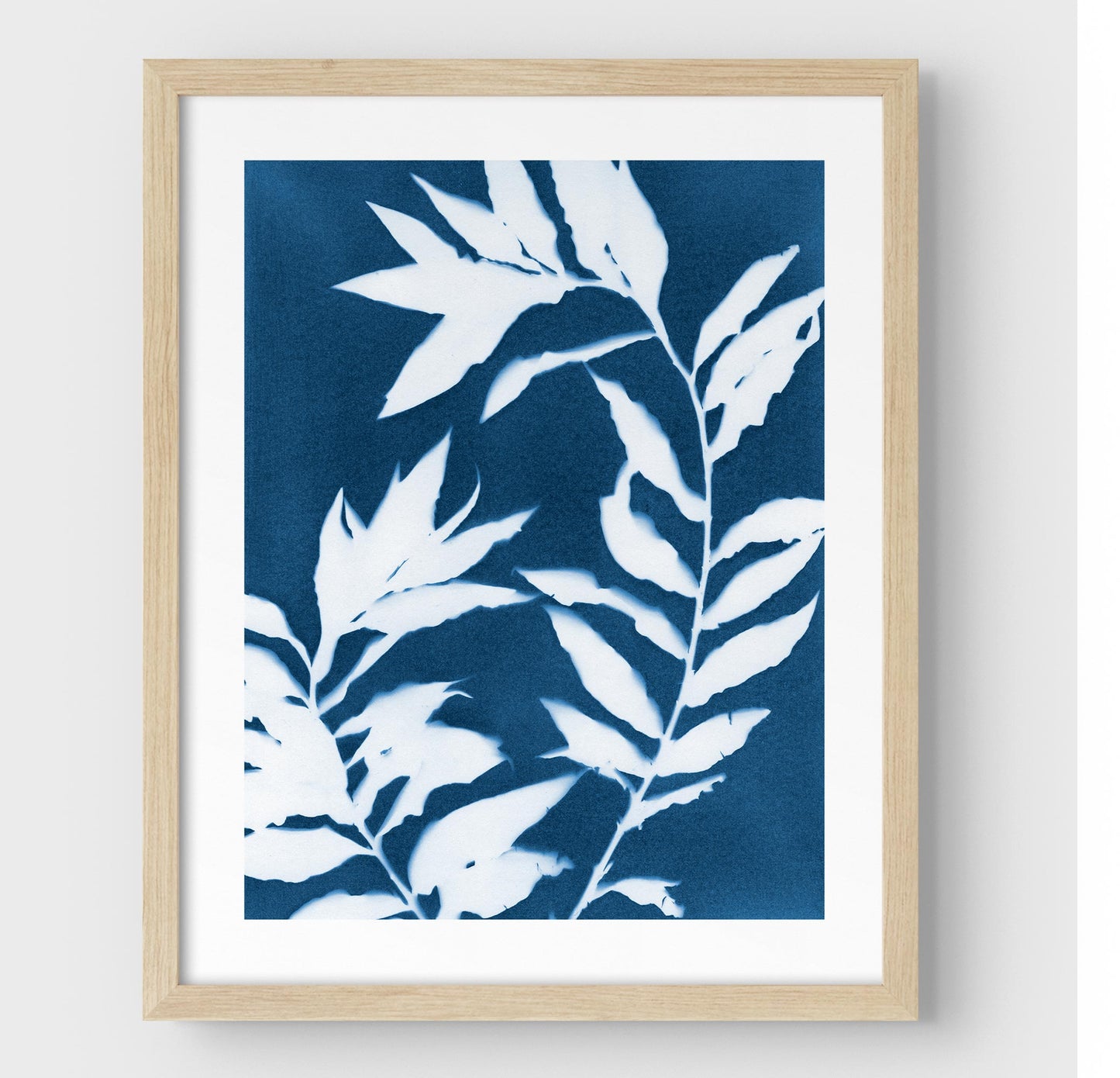 Framed cyanotype print featuring curved branches with pointed leaves in white against a deep blue background, enclosed in a natural wood frame with a white border.