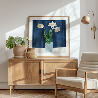 A cozy interior showcasing a framed square artwork of three white daffodils in a light blue vase against a dark blue background, displayed on a wooden credenza. The setting includes a potted monstera plant, a woven basket, and a beige armchair.