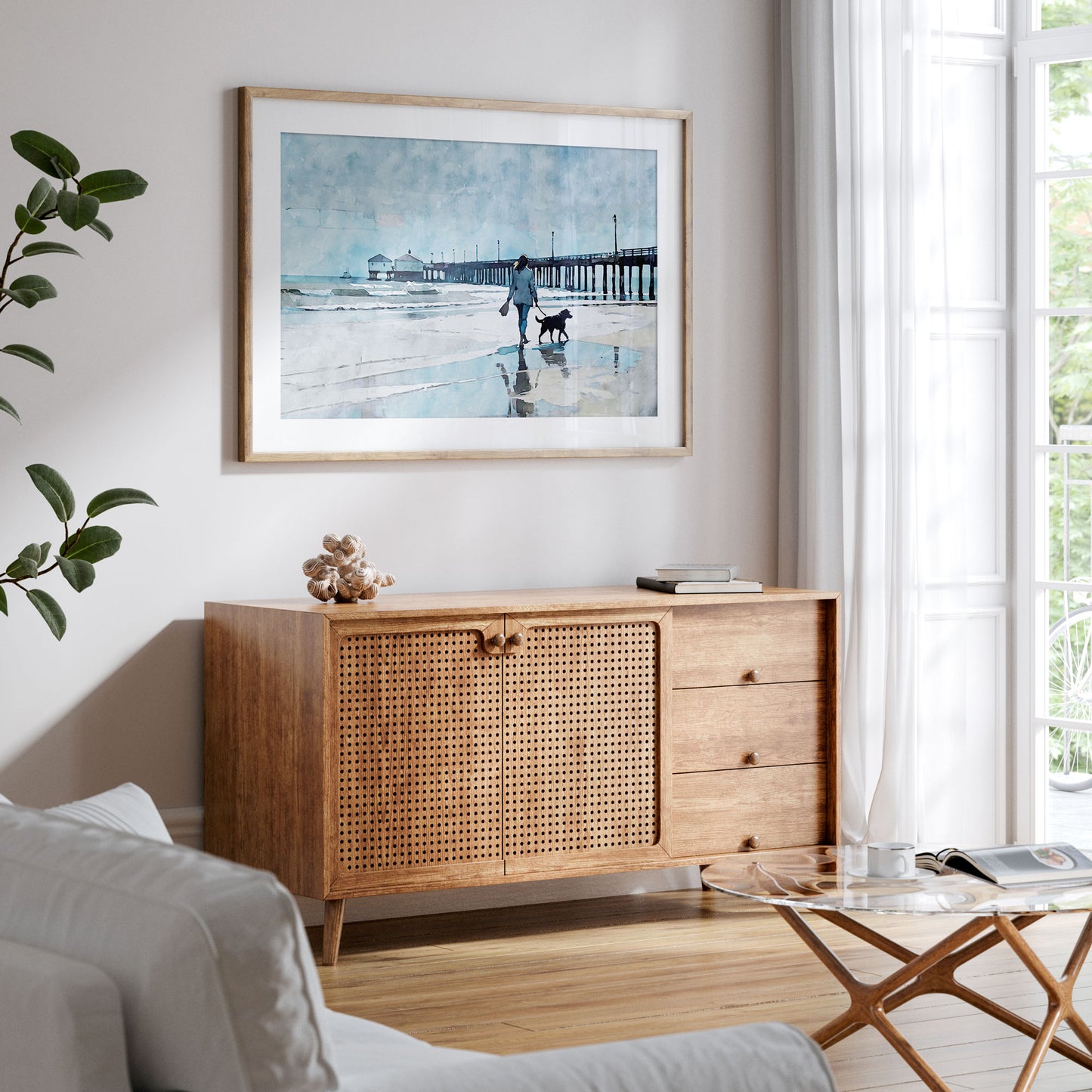 Framed artwork of a woman walking a dog on the beach hangs above a mid-century modern wooden cabinet in a bright living room with natural light.