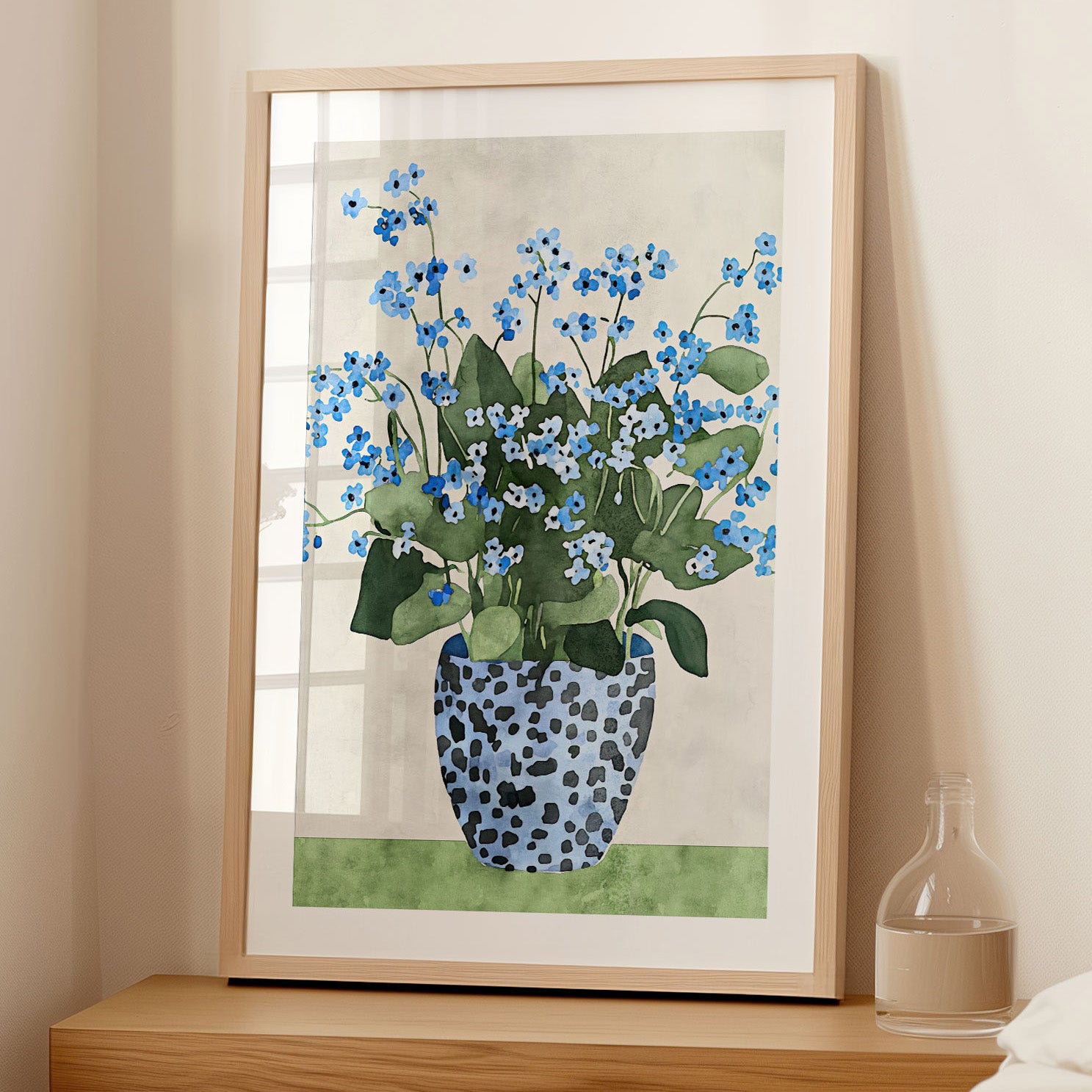 Framed artwork featuring a watercolor-style depiction of a bouquet of blue forget-me-not flowers with green leaves, arranged in a patterned blue vase, leaning against a wall on a wooden surface in a softly lit room.