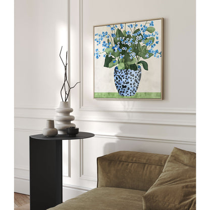 Framed artwork featuring blue forget-me-not flowers in a patterned blue vase hanging on a wall above a black side table with sculptural ceramic vases, next to an olive-green sofa in a softly lit, elegant interior.