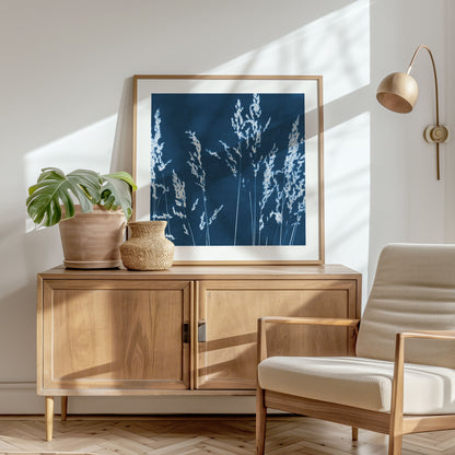 Framed cyanotype print of wild grasses on a wooden sideboard, featuring white botanical silhouettes on a deep blue background in a sunlit modern interior.