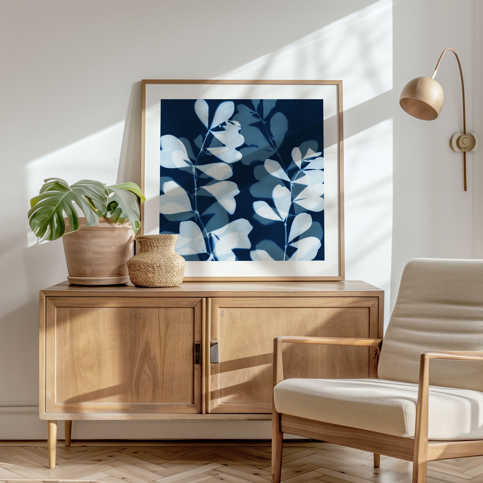 Framed cyanotype-style print resting on a wooden sideboard, showing layered white botanical silhouettes against a deep blue background, styled beside a terracotta planter and natural fiber vase in a sunlit modern interior.