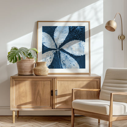 Framed cyanotype print of a five-lobed leaf with water droplets, displayed on a wooden sideboard in a sunlit room with neutral decor and modern furnishings.