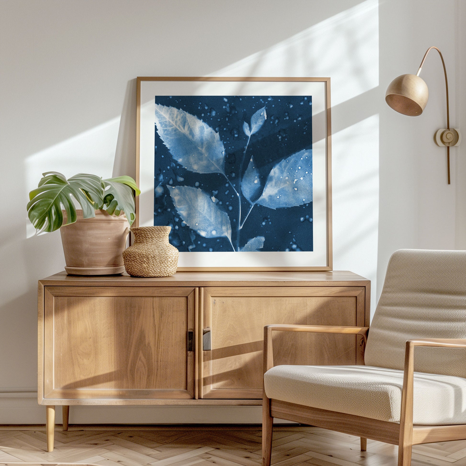 Framed cyanotype print of a leafy branch with water droplets, displayed on a wooden sideboard in a sunlit room with neutral decor and modern furnishings.