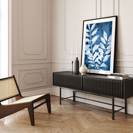 Framed cyanotype print of leafy branches in deep blue and white, leaning against a wall on a black sideboard in an elegant interior.