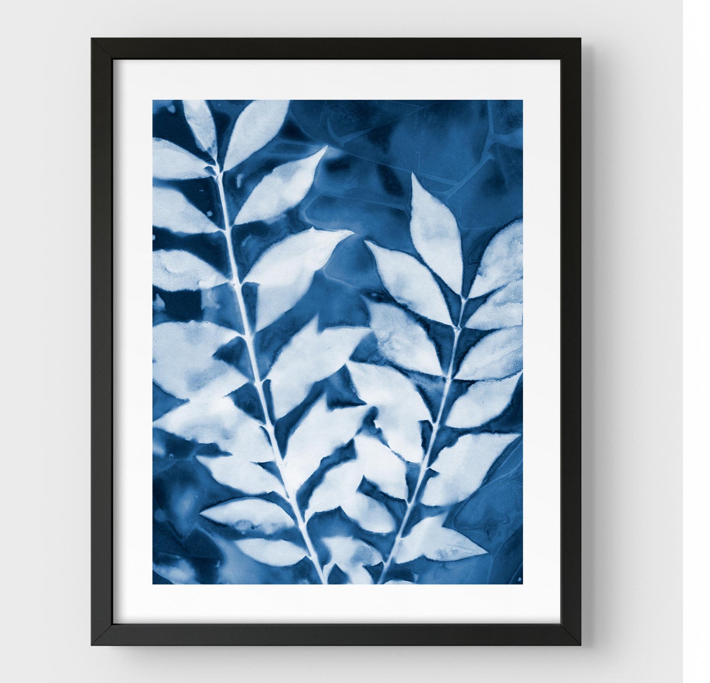 Framed cyanotype print featuring two leafy branches in deep blue and white, encased in a black frame with a white border.