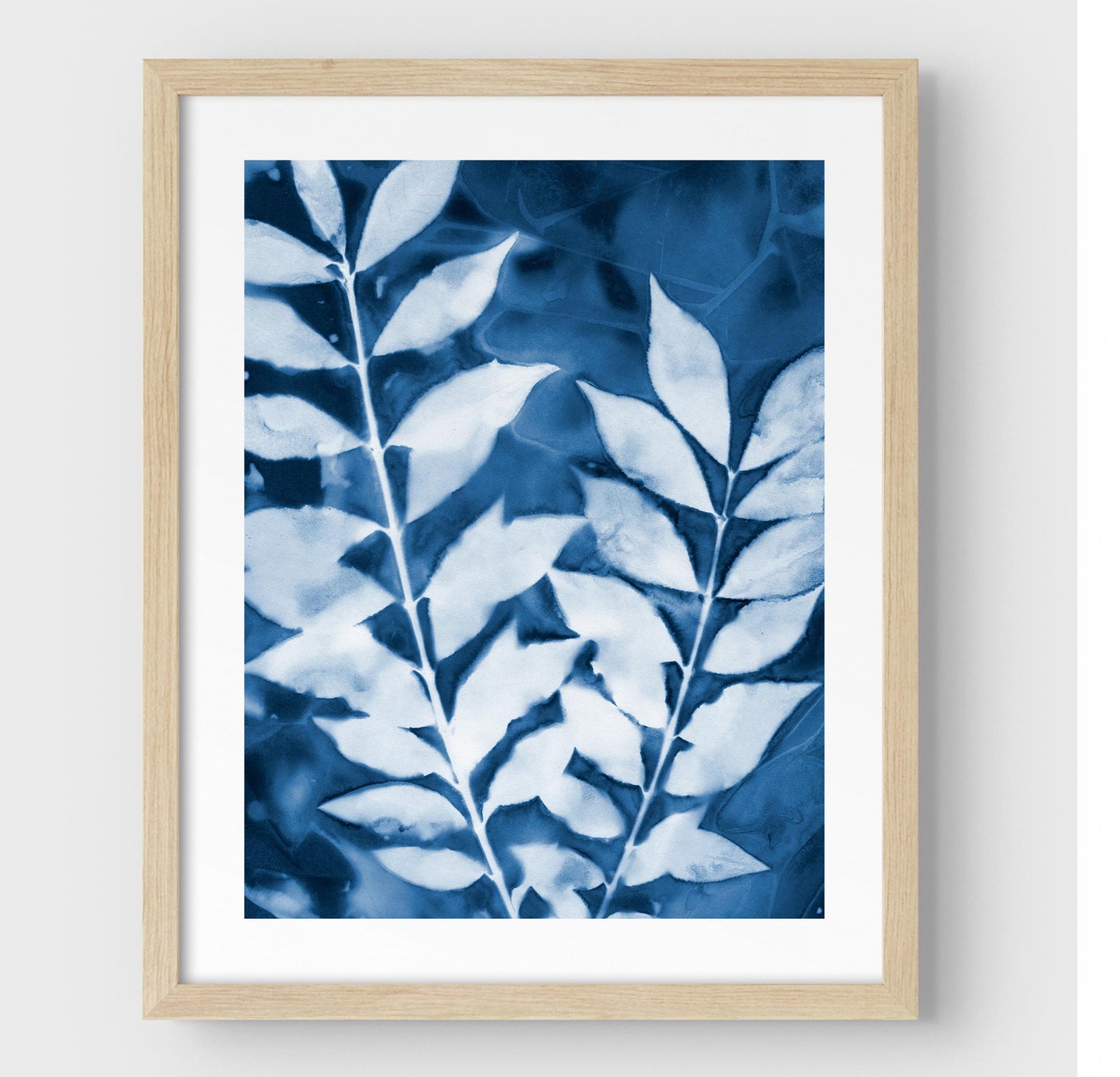 Framed cyanotype print featuring two leafy branches in deep blue and white, encased in a natural wood frame with a white border.