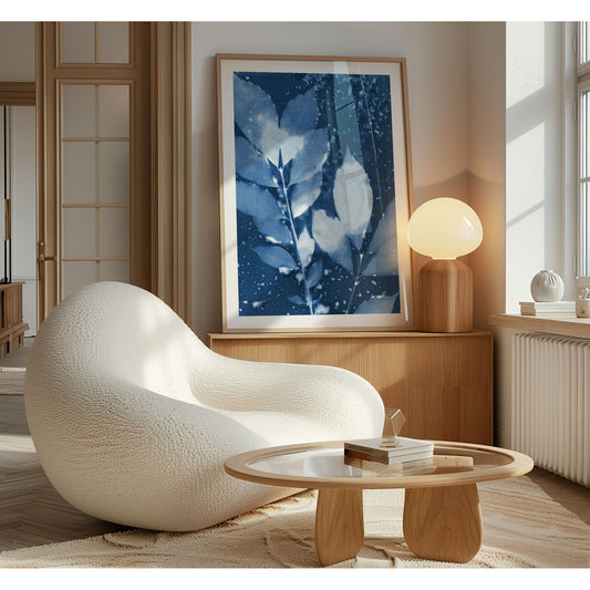 Framed cyanotype print of delicate leaves in deep blue and white tones, leaning against a wooden sideboard in a softly lit modern living space.
