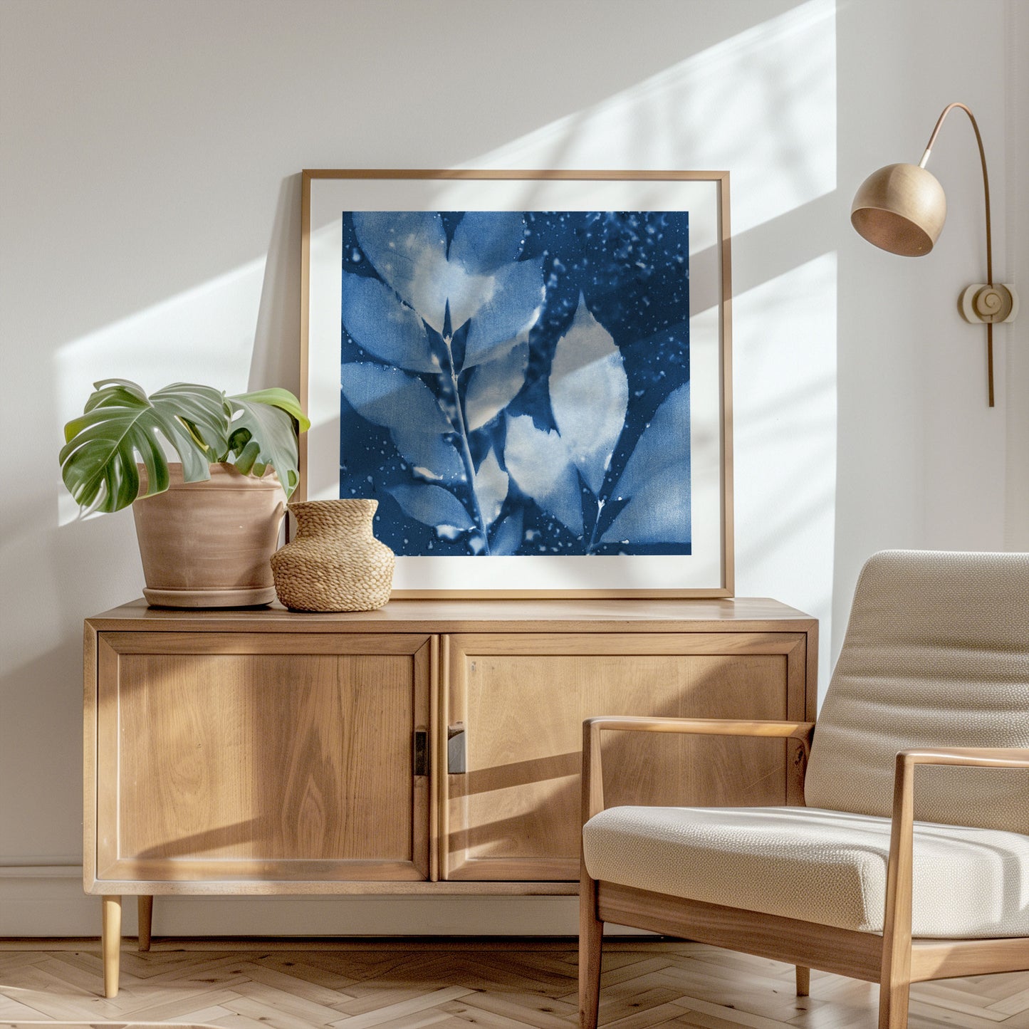 Framed cyanotype print of abstract leaves in deep blue and white, displayed on a wooden sideboard in a sunlit room with neutral decor and natural accents.