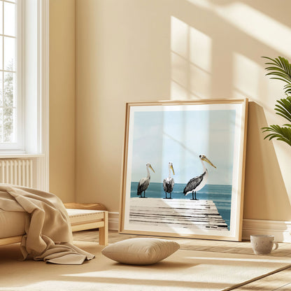 Framed painting of three pelicans on a dock by the ocean, leaning against a wall in a softly sunlit, neutral-toned room with cozy furnishings and natural textures.